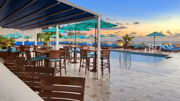 A poolside area with lounge chairs and umbrellas, a pool, and a view of the ocean at sunset.