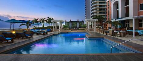 Zona de piscina con sillas de descanso y sombrillas, rodeada por un edificio de gran altura con balcones.
