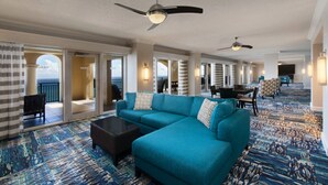 Un hall d’hôtel spacieux, avec un grand canapé turquoise, une table basse et une moquette à motifs. On y trouve des ventilateurs de plafond, un coin repas avec des tables et des chaises, et une vue à travers les portes vitrées.