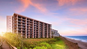 Un hotel de varios pisos con balcones, situado cerca de una playa bajo un cielo despejado.