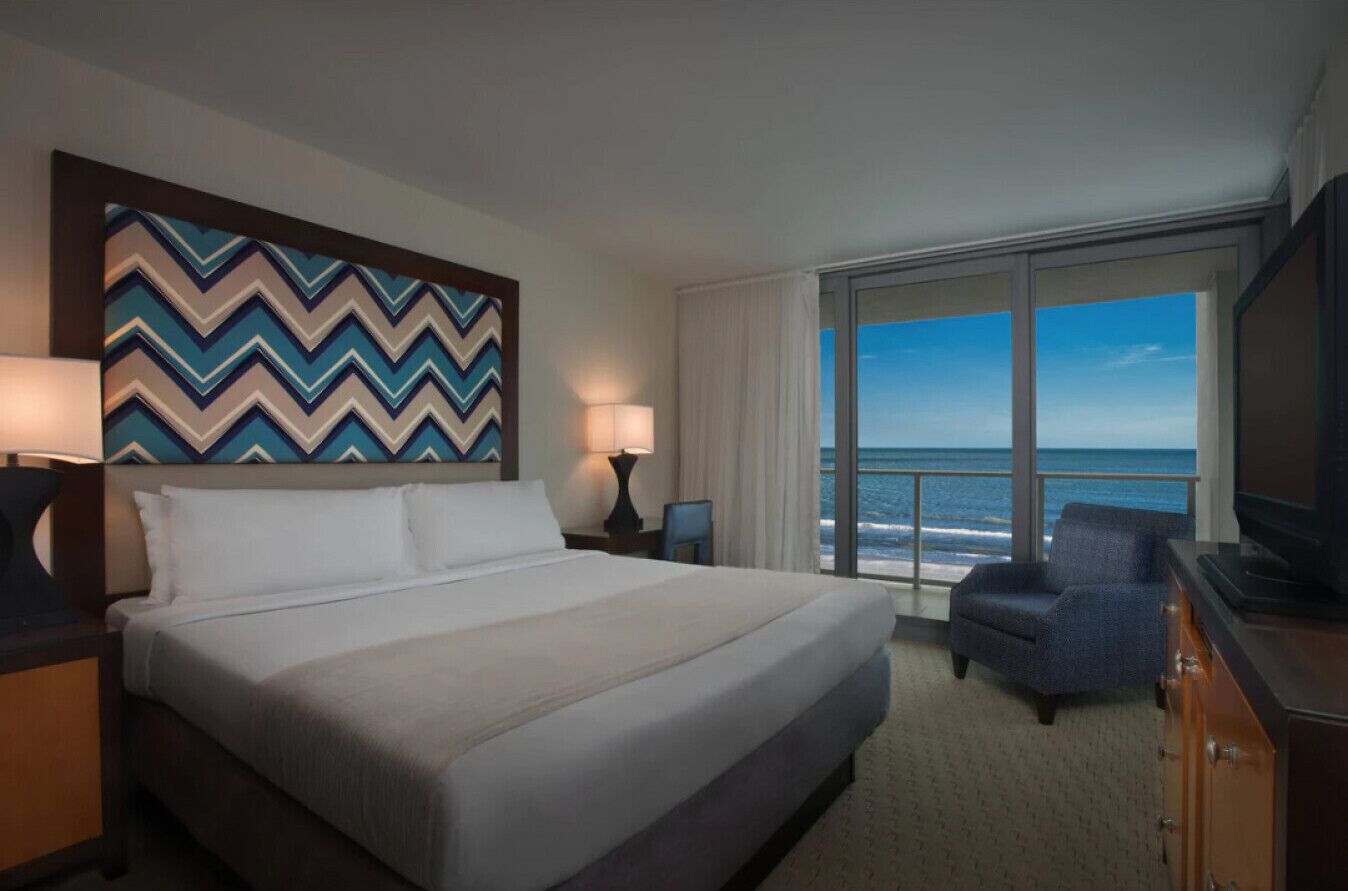 A hotel room with a large bed, a television, a chair, and a view of the ocean.
