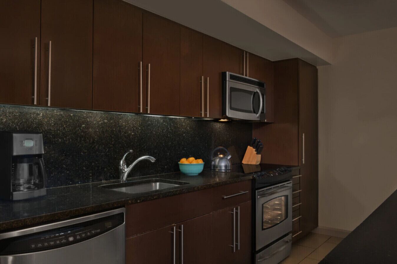 A modern kitchen with dark wood cabinets, a stainless steel dishwasher, a built-in oven, a microwave, and a granite countertop.