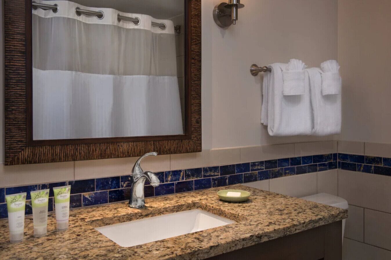 Un baño con encimera de granito, un lavabo, un espejo, un perchero para toallas y una botella de loción.