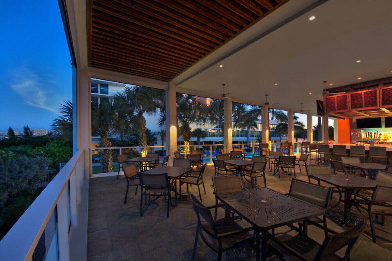 An outdoor patio area with tables and chairs, large windows, and a pool in the background.