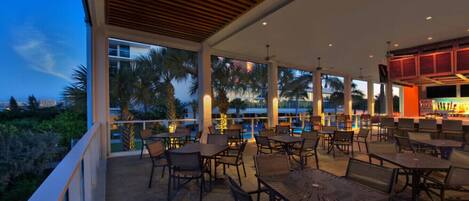 An outdoor patio area with tables and chairs, large windows, and a pool in the background.