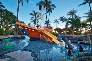 Explore the fun-filled water play area featuring a ship-shaped slide, pool with water jets, and a small waterfall surrounded by palm trees.