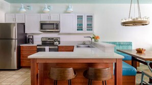 Cocina moderna con armarios blancos, un refrigerador de acero inoxidable, un microondas y una cafetera. Hay una isla de madera con dos taburetes y un banco tapizado azul en la mesa de comedor.