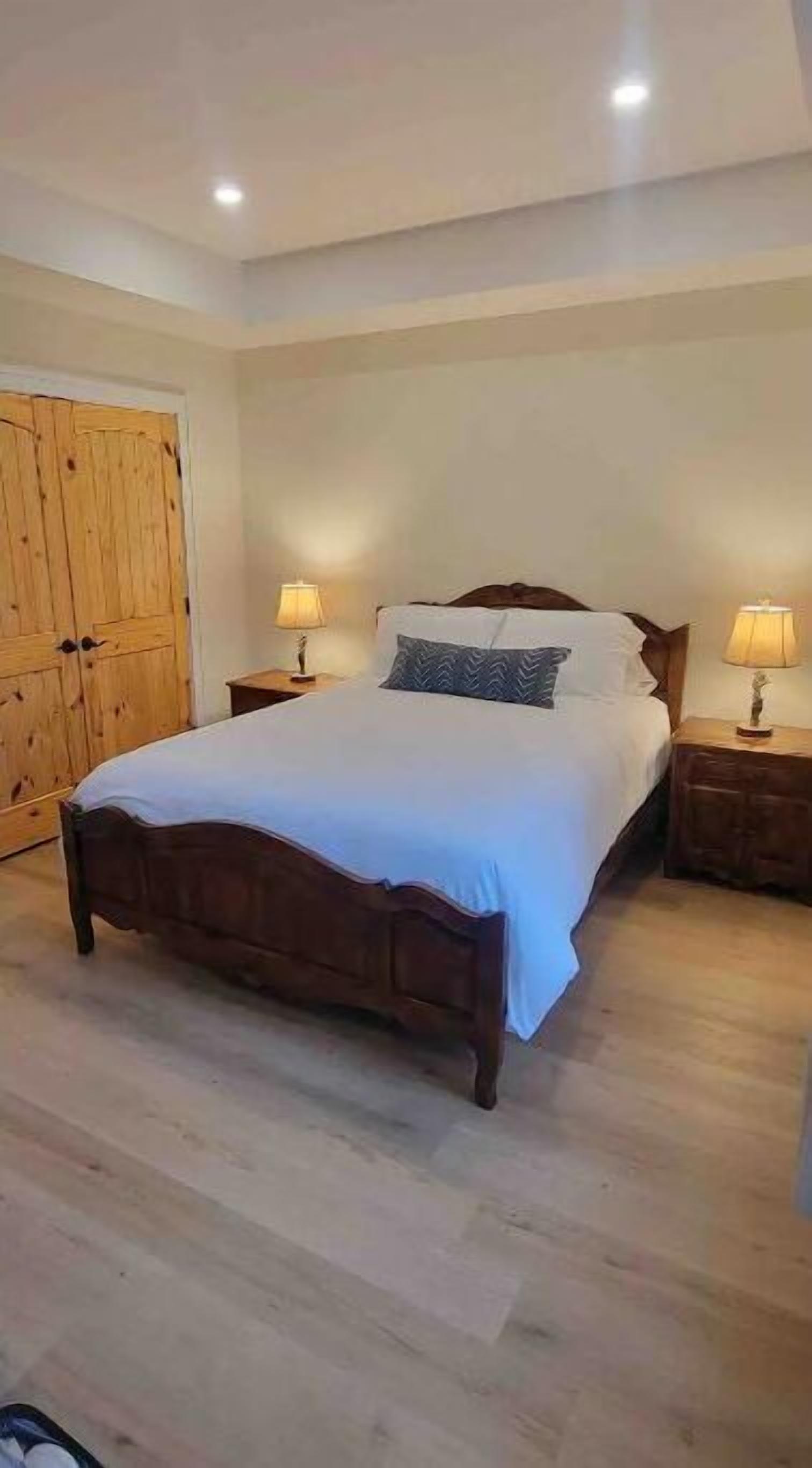 A bedroom with a wooden bed, two bedside tables with lamps, a wooden wardrobe, and a light-colored floor.