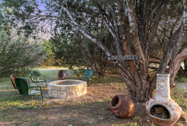 A rustic outdoor setting with a stone fire pit, green metal chairs, and a large tree.