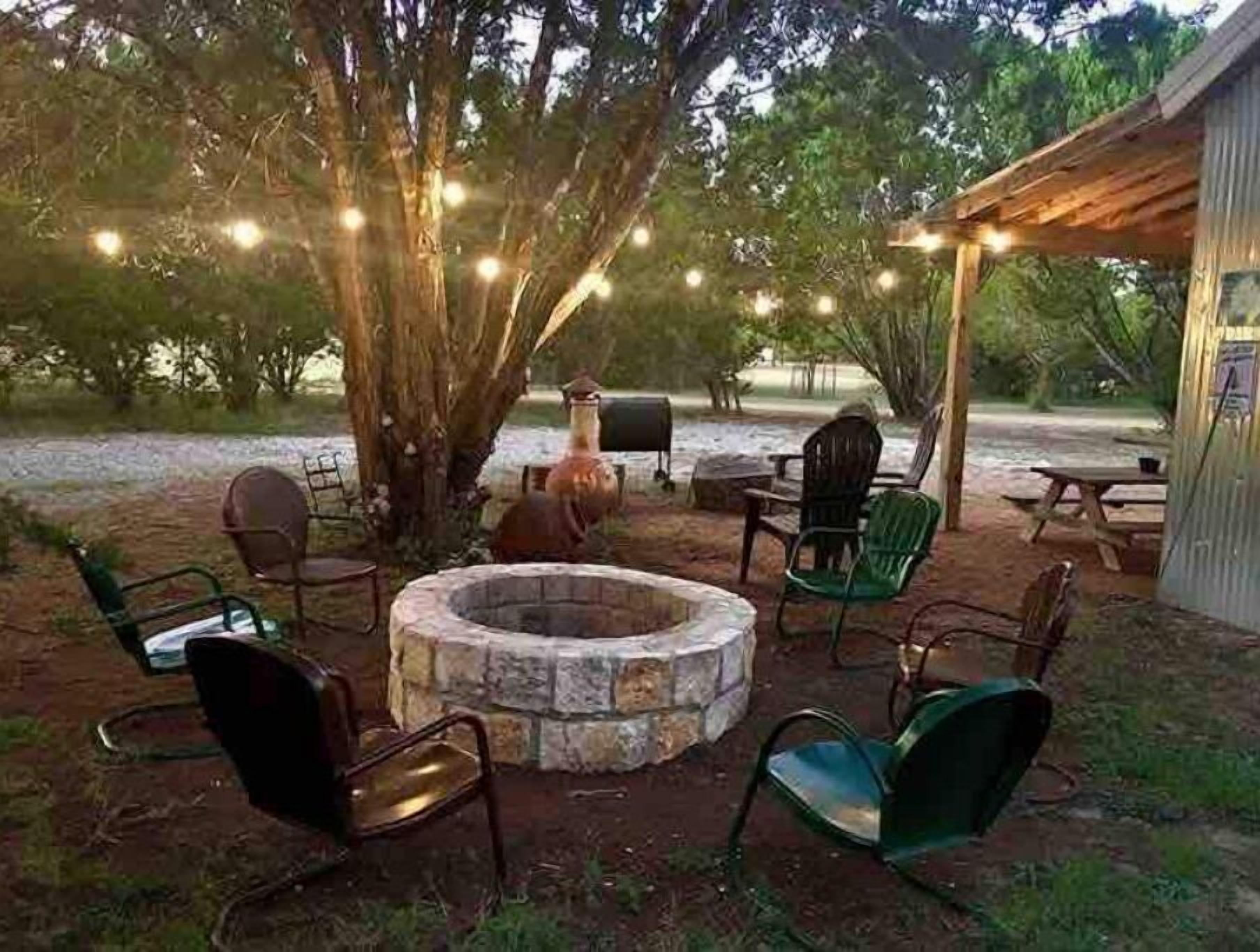 Area salotto esterna con un braciere in pietra, circondata da sedie e tavoli, sotto un albero illuminato da luci decorative.