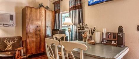 A rustic dining area with a wooden table, white chairs, a vintage coffee maker, and a television on the wall.