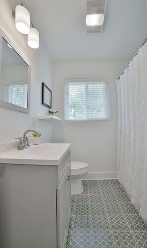 Ein Badezimmer mit Waschbecken, Spiegel und Toilette. Ein Fenster mit Jalousien und eine Duschvorhang sind vorhanden.