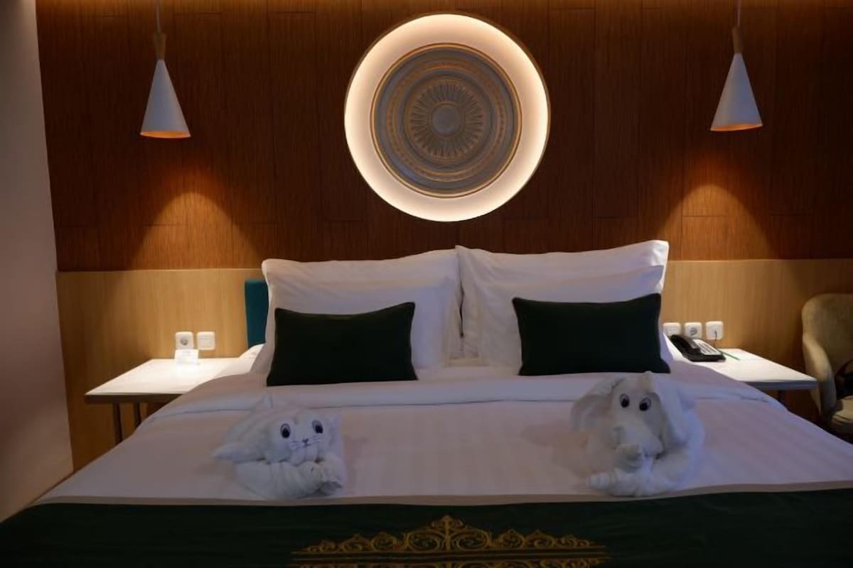 A hotel room with a bed, two pillows, a bedside table, a lamp, and a wall-mounted light fixture.