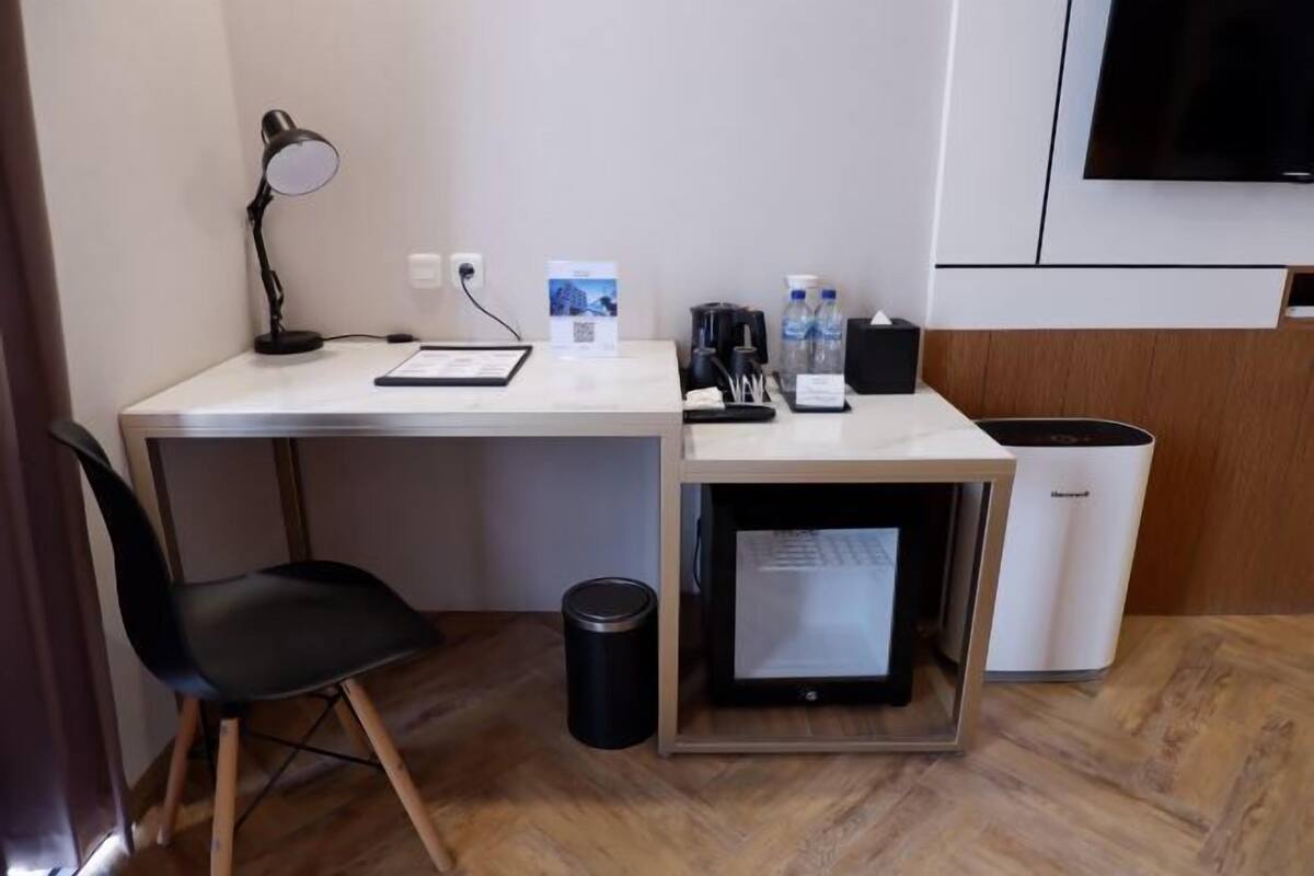 A compact hotel room with a desk, chair, desk lamp, refrigerator, trash bin, and a television on the wall.