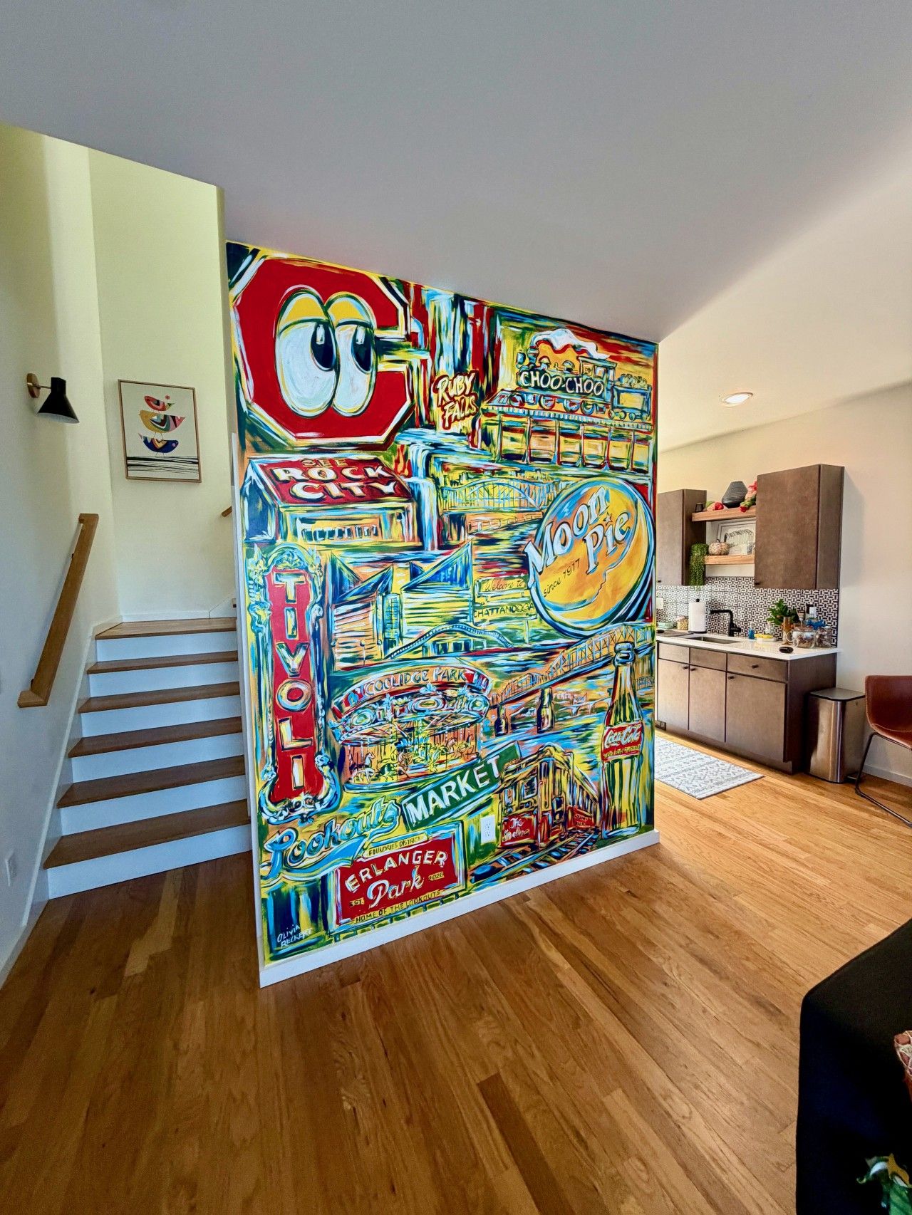 Un mural vibrante y colorido en un pasillo, con una escalera y una zona de cocina al fondo.