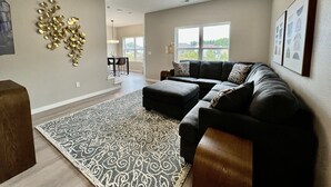 A spacious living room with a dark-colored sectional sofa, a patterned rug, and a dining area with a table and chairs. - New Construction Townhome - 3 King Beds/2.5 Bathroom! (Cedar Falls)