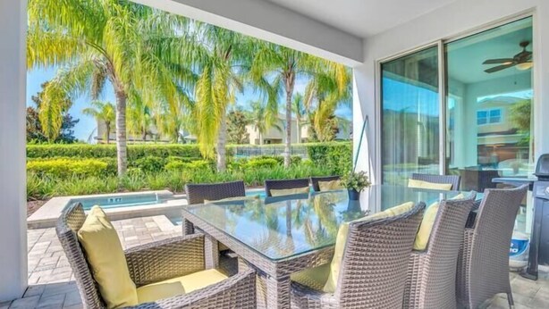 A modern outdoor dining area with wicker furniture and a glass table, overlooking a pool and lush greenery. - Spacious Pool Home Close To Disney (Kissimmee)