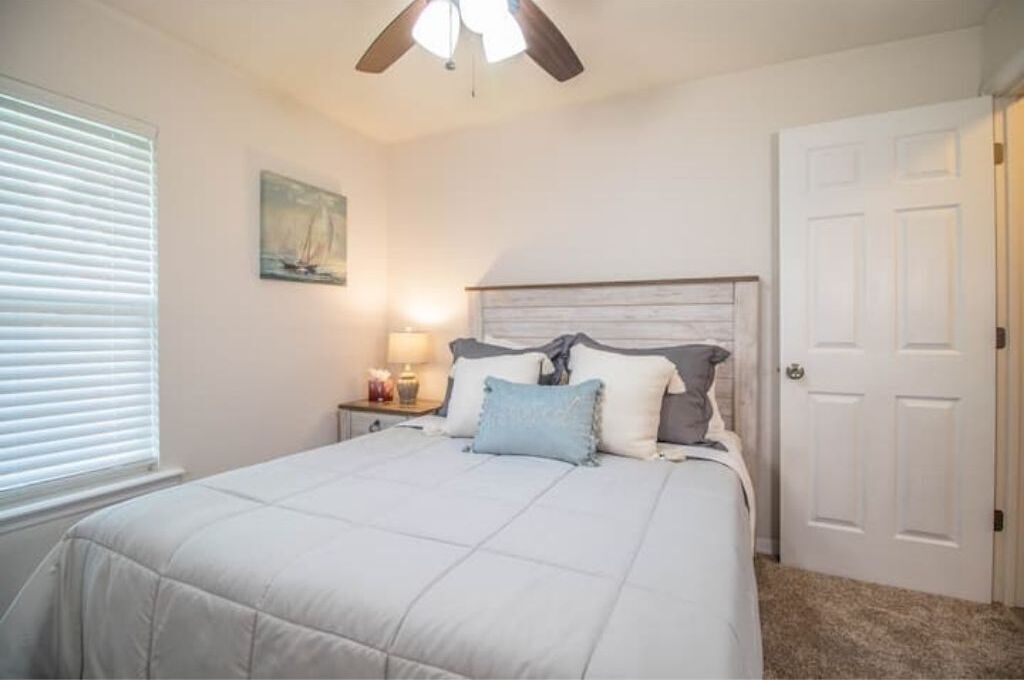 Une chambre à coucher comprenant un lit, une table de chevet, un ventilateur de plafond, une porte et une fenêtre avec des stores.