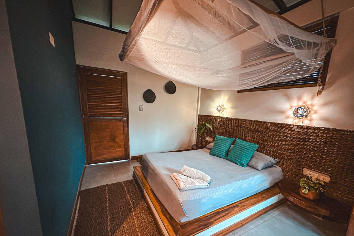 Un dormitorio con cama, cabecera de madera, mesita de noche, planta en maceta y ventana con cortina.