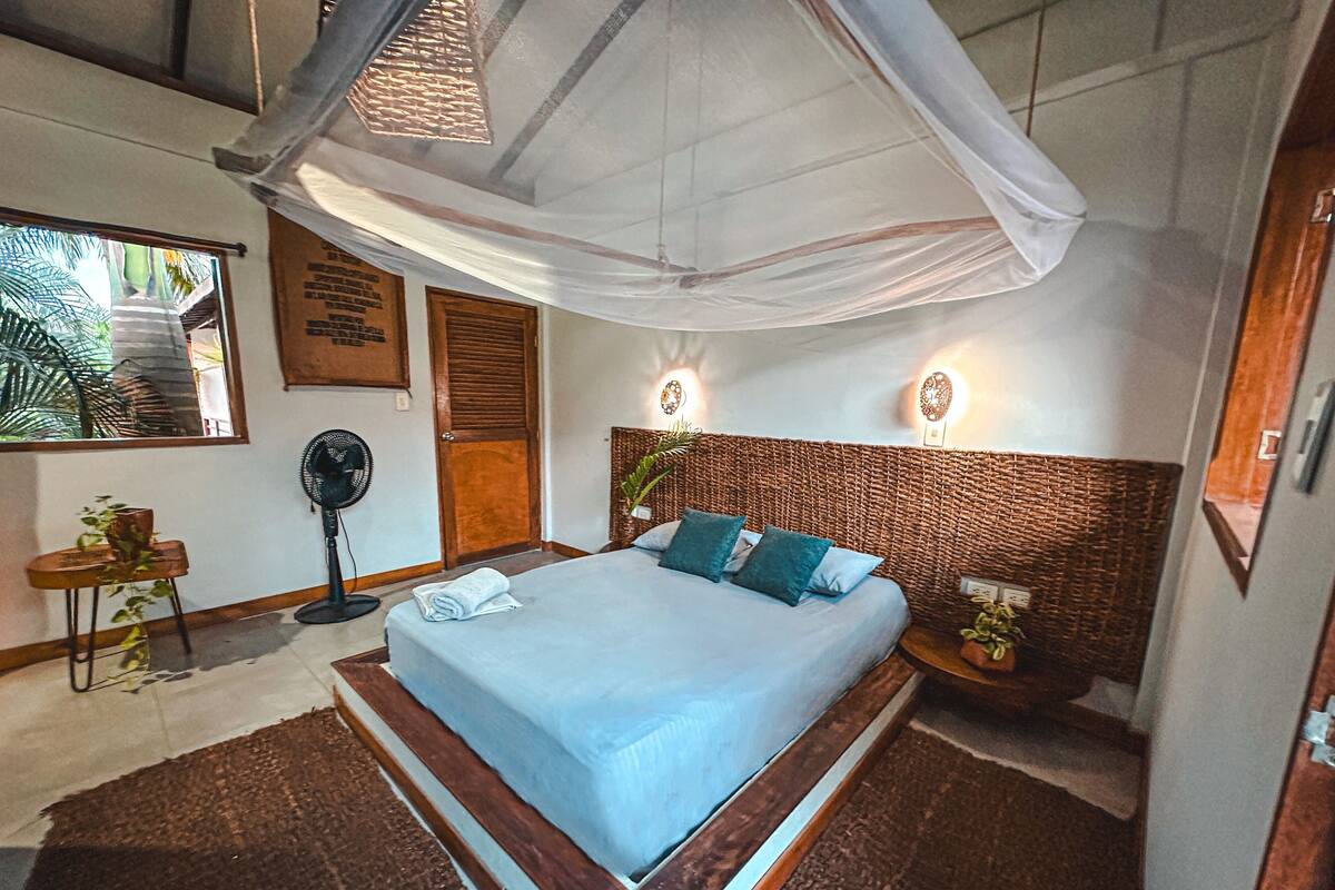 Un dormitorio con cama, ventilador, mesita de noche, luz de pared y ventana con vistas.