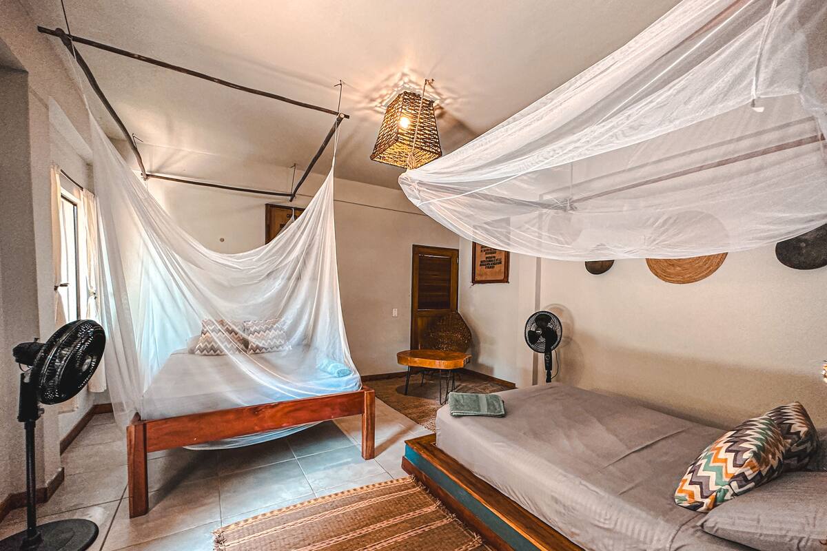 Un dormitorio con cama, una silla, un ventilador de pie, una mesita de noche y una lámpara colgante.