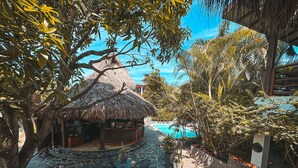 Uma cabana com telhado de palha, piscina e área de estar ao ar livre, rodeada por plantas e árvores tropicais.