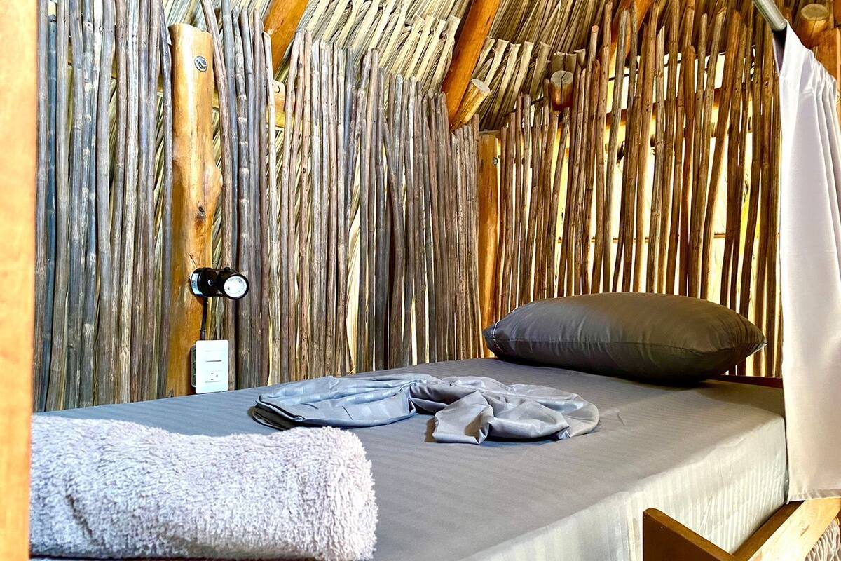 Una cama individual con un colchón gris, una toalla blanca y un almohadón gris en una habitación con pared de bambú.