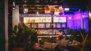 Uma área de estar acolhedora com móveis de vime, um bar com cardápio em lousa e diversas plantas.