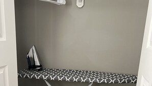 Une petite pièce avec un mur gris, une planche à repasser à motifs, une autre planche à repasser blanche, un étendoir et un petit panier à linge.