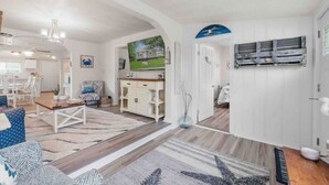 Un vaste salon comprenant un grand canapé, une table basse et un coin repas avec des chaises. Il y a également une cuisine avec des meubles blancs et une table à manger. La pièce est dotée d'un parquet et d'un grand tapis.
