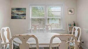 Une salle à manger avec une table ronde et des chaises blanches, de grandes fenêtres équipées de stores et un tableau accroché au mur.