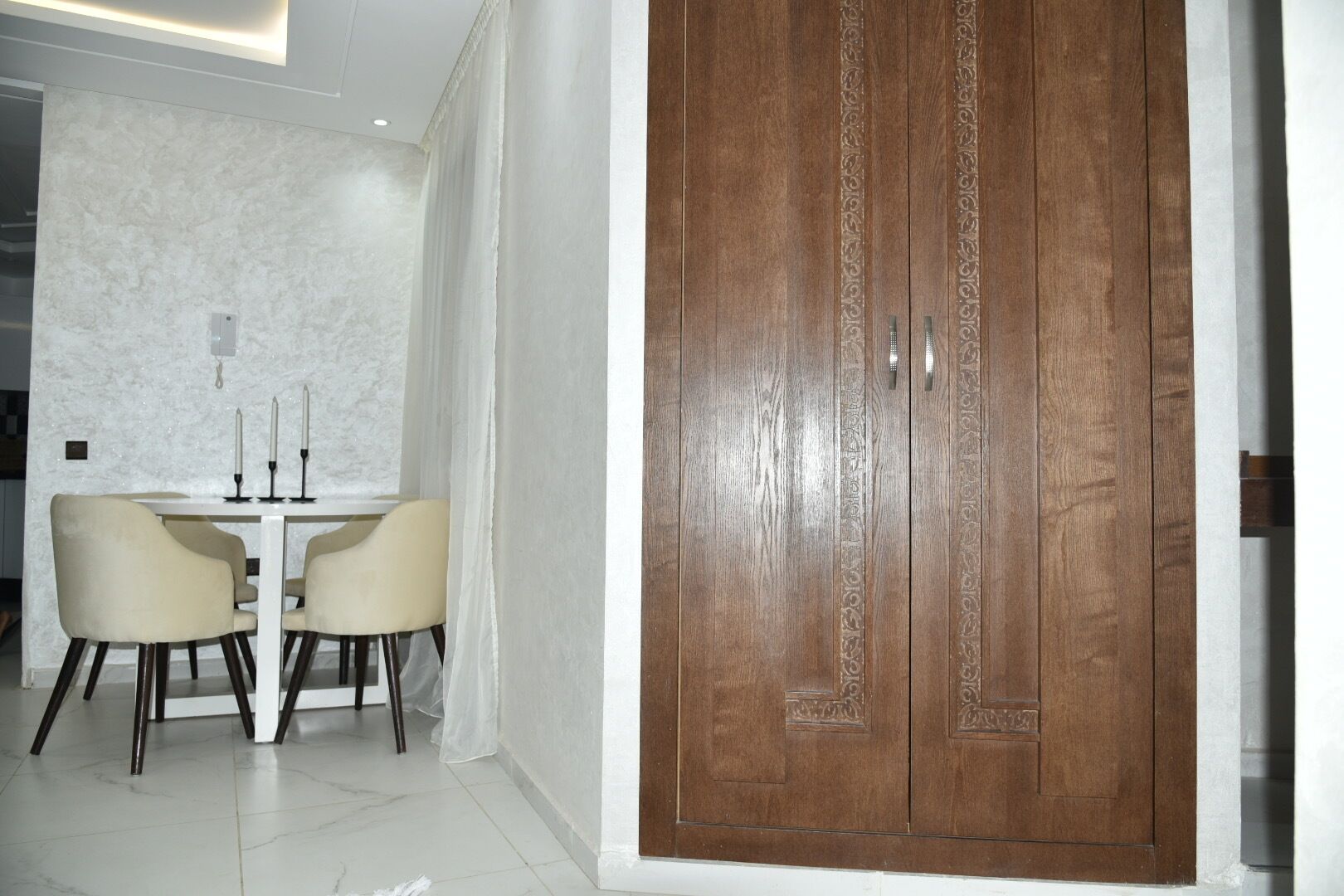 Un comedor con una mesa blanca y dos sillas, una puerta de madera con intrincados grabados y una pared blanca con textura.