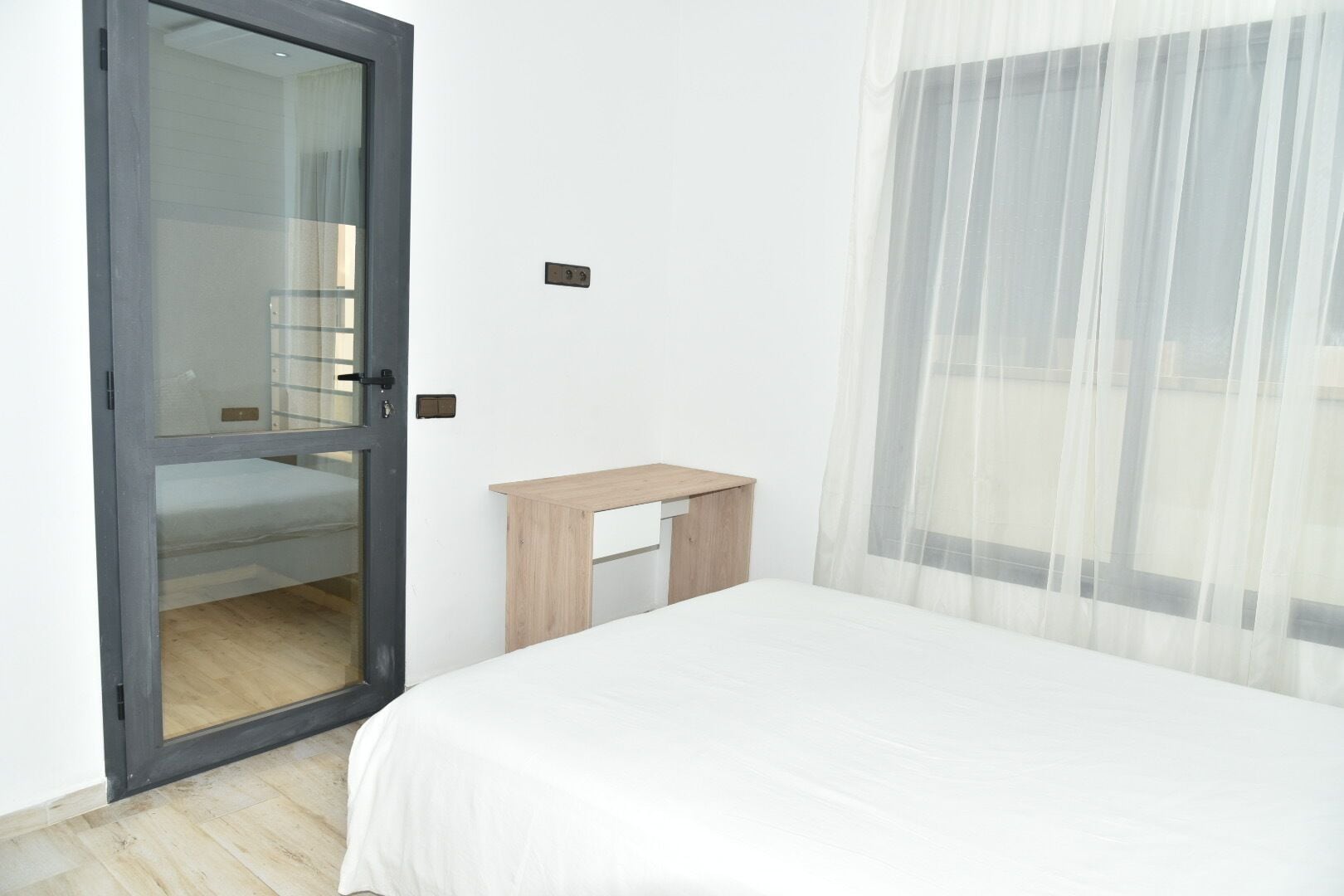 Un dormitorio con cama, una mesita de noche de madera, un ventanal corredero y vistas a través de cortinas translúcidas.