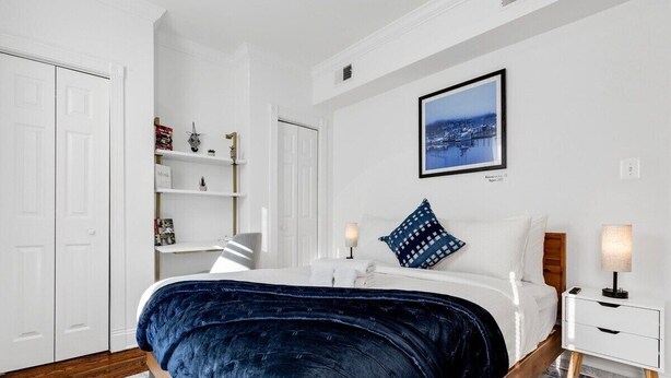A bedroom with a bed, bedside lamps, a nightstand, a shelf with books and decorative items, and a framed picture on the wall. - Entire Rowhome in Northwest Washington (Dedicated Parking Spot Available) (Washington)