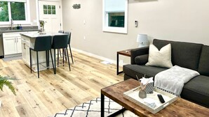 A modern living room with a dark grey sofa, a wooden coffee table, and a kitchen area with a bar counter and two black chairs. - 5 min to DWTN, BART & Park| Private Deck w/ View (Concord)