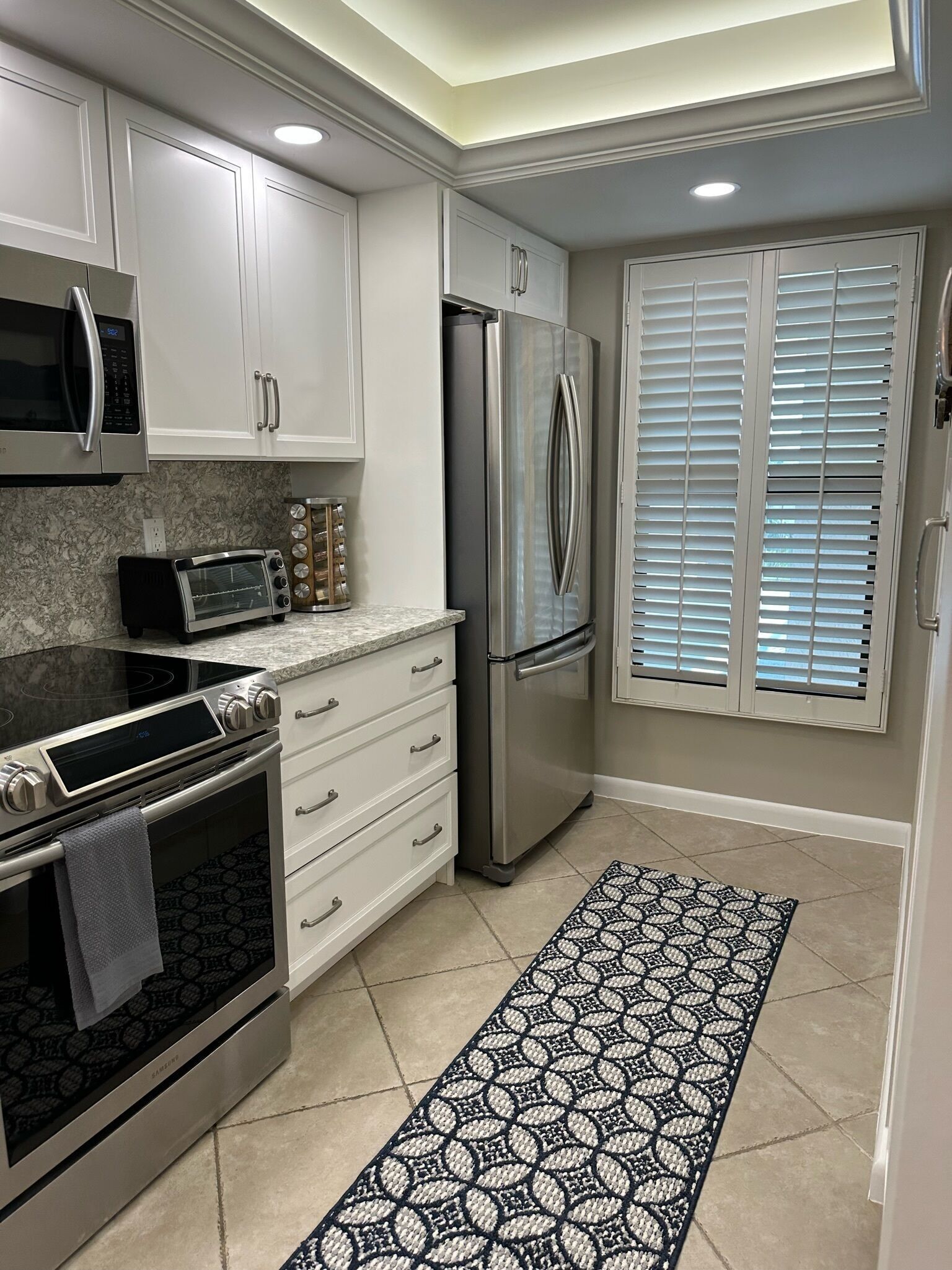 Una cocina moderna con armarios blancos, electrodomésticos de acero inoxidable, un tapete estampado y una ventana con persianas.