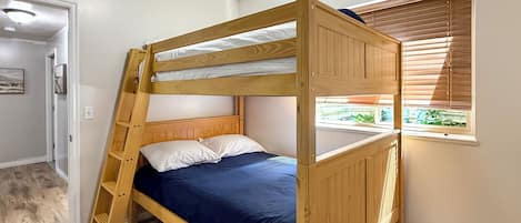 Una cama litera con escalera de madera, una ventana con persianas y una puerta que da a otra habitación.
