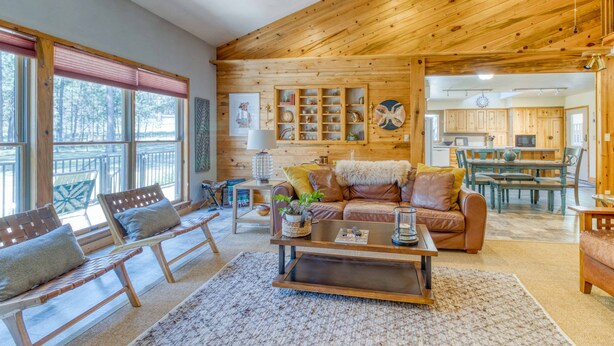 A spacious living room with a wooden ceiling, a large window with blinds, a sofa with cushions, a coffee table, and a kitchen area in the background. - 1-acre retreat with firepits, wraparound porch & a relaxing 6-person hot tub (Sisters)