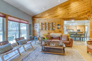 A spacious living room with a wooden ceiling, a large window with blinds, a sofa with cushions, a coffee table, and a kitchen area in the background.