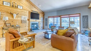 A cozy living room with a fireplace, wooden walls, and a large window. - 1-acre retreat with firepits, wraparound porch & a relaxing 6-person hot tub (Sisters)