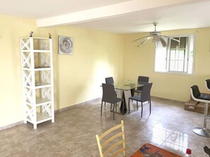 A dining area with a glass table and four chairs, a white shelving unit, a ceiling fan, and a window with blinds.