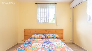 A bedroom with a wooden bed, colorful floral bedding, a window with a white curtain, and an air conditioning unit on the wall.