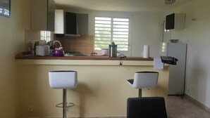 A compact kitchen with a gas stove, a refrigerator, and a window with blinds. There are two bar stools and a small countertop area. - Joss plein  air (Le Gosier)