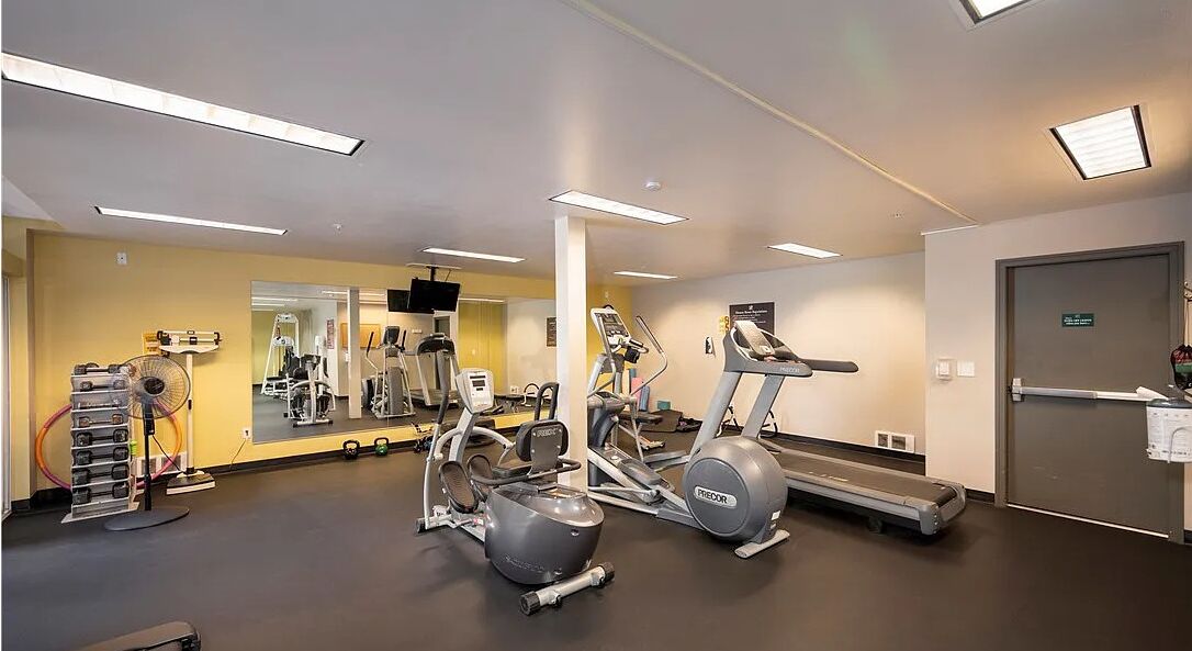 Gimnasio con máquinas de cardio, equipamiento de pesas y un espejo grande.