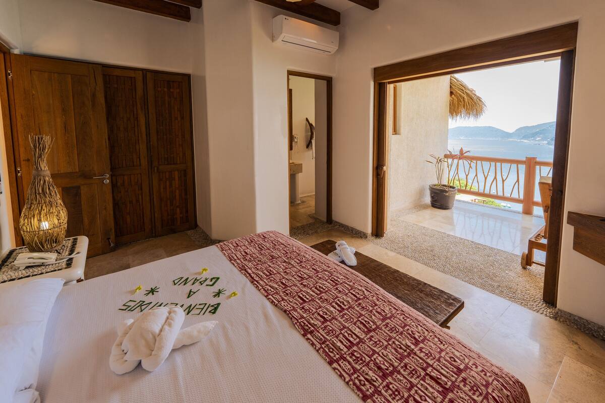 Une chambre d’hôtel avec un lit, une porte en bois, un miroir et un balcon donnant sur la mer.