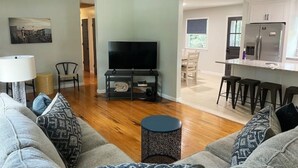 A living room with a TV, sofa, and coffee table. There is a kitchen area with a refrigerator and bar stools.