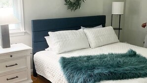 A neatly made bed with a white quilt and a teal throw blanket, a white bedside table with drawers, a dark blue headboard, a wall-mounted lamp, and a wreath on the wall.