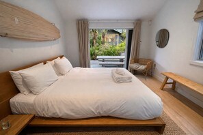 A bedroom with a bed, a bench, a chair, and a view of the outdoors. - Serenity - Palm Beach Pathway Retreat (Waiheke Island)