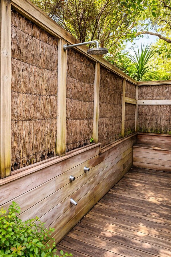 A wooden deck with a built-in shower area, surrounded by a thatched wall and greenery. - Serenity - Palm Beach Pathway Retreat (Waiheke Island)