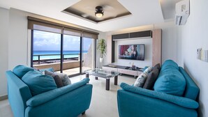 Ein modernes Wohnzimmer mit großem Fernseher, zwei blauen Sofas, einem Glastisch und Meerblick.